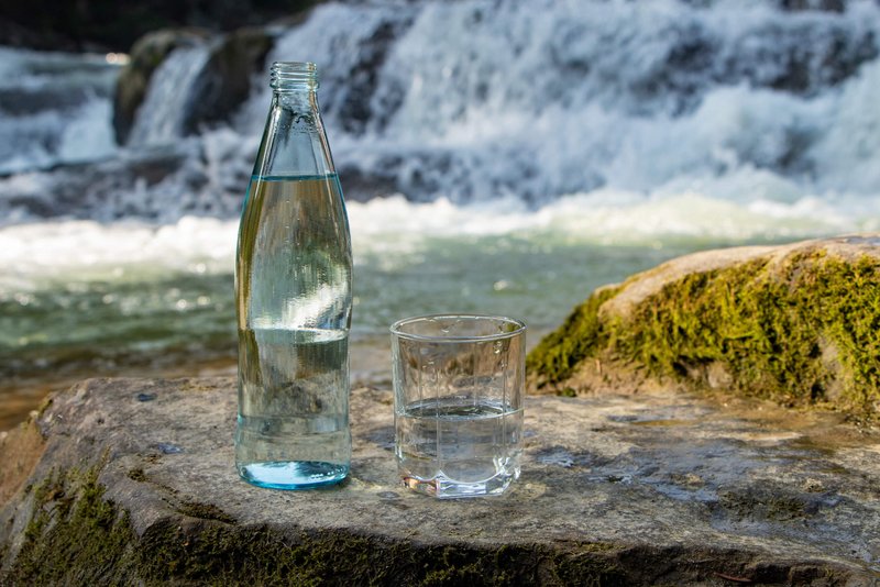 Natürliches Mineralwasser in einer Glasflasche und einem Trinkglas auf einem großen Stein. Dahinter ein Fluss.